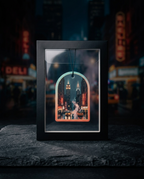 A black shadow box frame containing a miniature New York City street scene with illuminated skyscrapers and traffic, displayed on a dark stone surface with a blurred city night background.