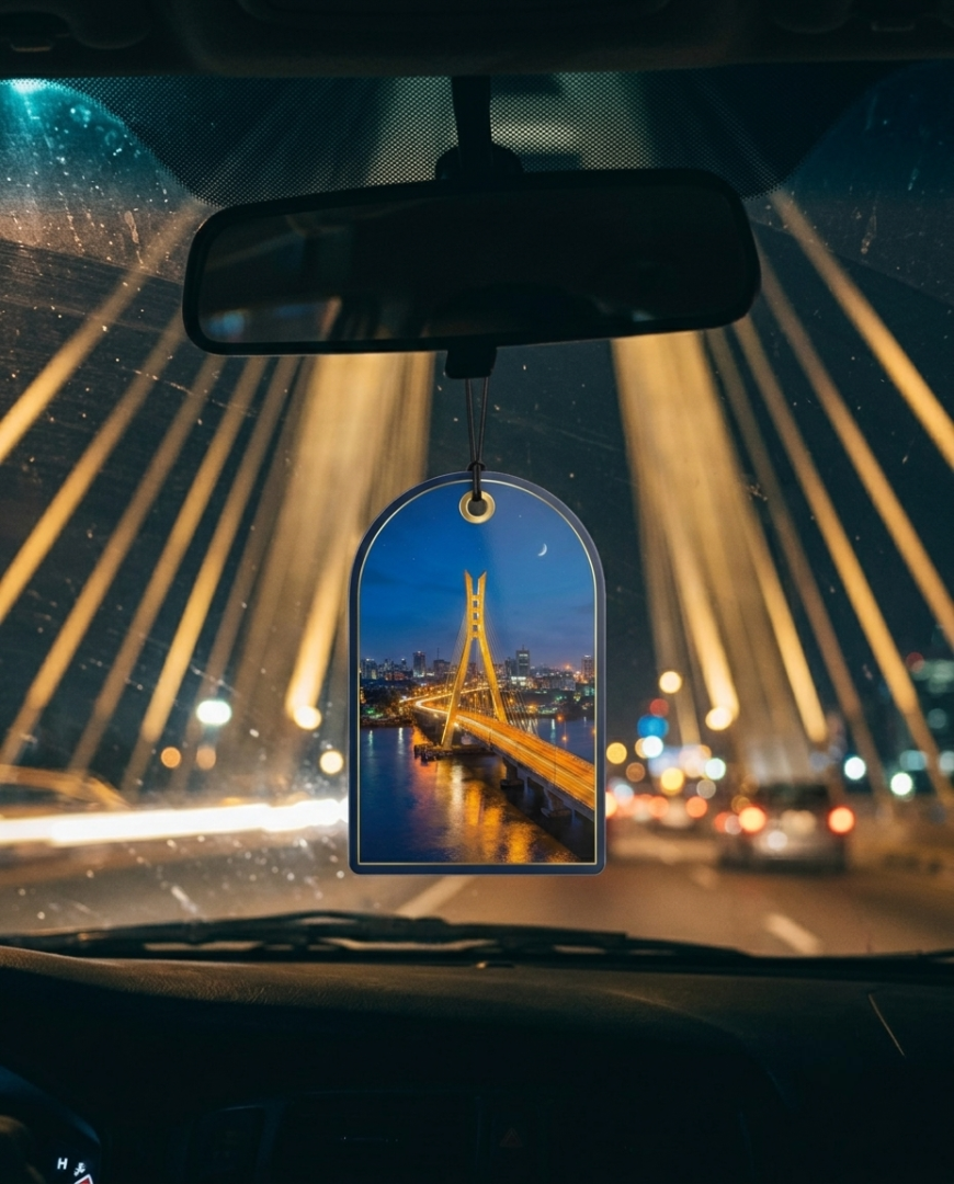 An arched car air freshener hanging from a rearview mirror inside a car, featuring a printed image of a illuminated bridge at night over water.