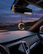 A gold metallic letter 'C' car air freshener or ornament hanging from a rearview mirror inside a car cabin at dusk.