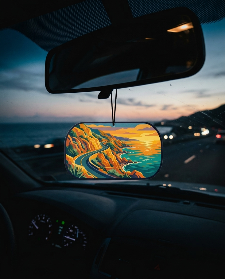 A colorful landscape car air freshener hanging from a rearview mirror in a car at dusk, showing a stylized coastal road scene in yellow, orange, and blue-green tones.