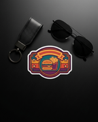 A colorful retro-style sticker featuring a hamburger and french fries design, displayed on a dark gray background next to a black leather keychain and black sunglasses.