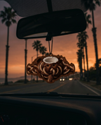 A carved wooden car air freshener shaped like a coiled snake or dragon, hanging from a car's rearview mirror during a sunset drive with palm trees in the background.