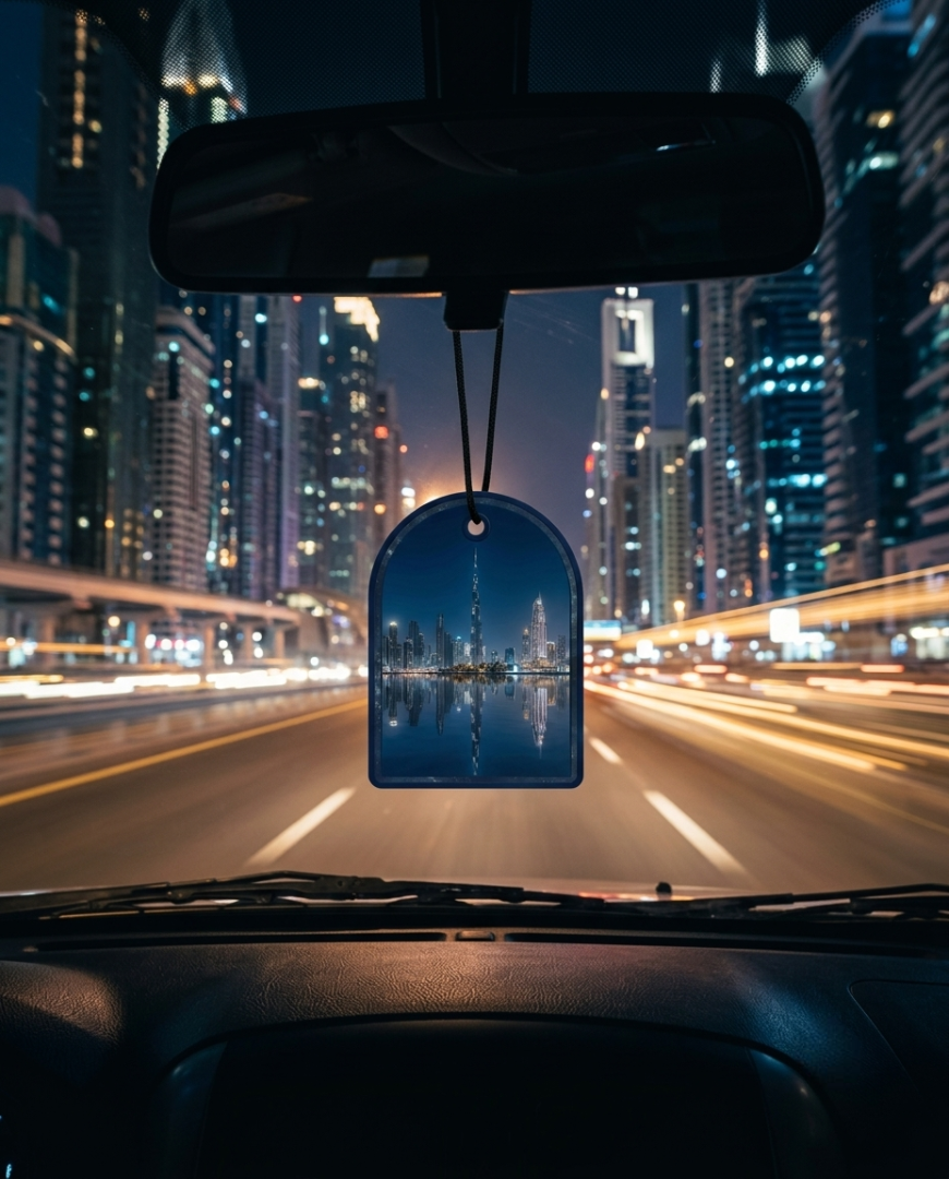 A car air freshener hanging from the rearview mirror in a car, showing a printed city skyline reflection at night with light trails from streetlights.