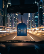 A car air freshener hanging from the rearview mirror in a car, showing a printed city skyline reflection at night with light trails from streetlights.
