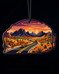 A die-cut hanging ornament featuring a stylized desert landscape with a winding road, cars, cacti, mountains, and an orange sunset sky, shown against a black background.