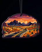 A die-cut hanging ornament featuring a stylized desert landscape with a winding road, cars, cacti, mountains, and an orange sunset sky, shown against a black background.