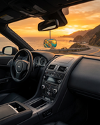 A car interior with a hanging round air freshener featuring a colorful coastal landscape print, shown from the driver's seat with a winding coastal road and ocean at sunset visible through the windshield.
