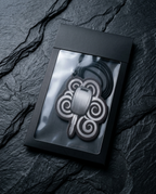 A black metal keychain with a traditional Celtic knot design, presented inside a black gift box with a clear window, set against a dark slate background.