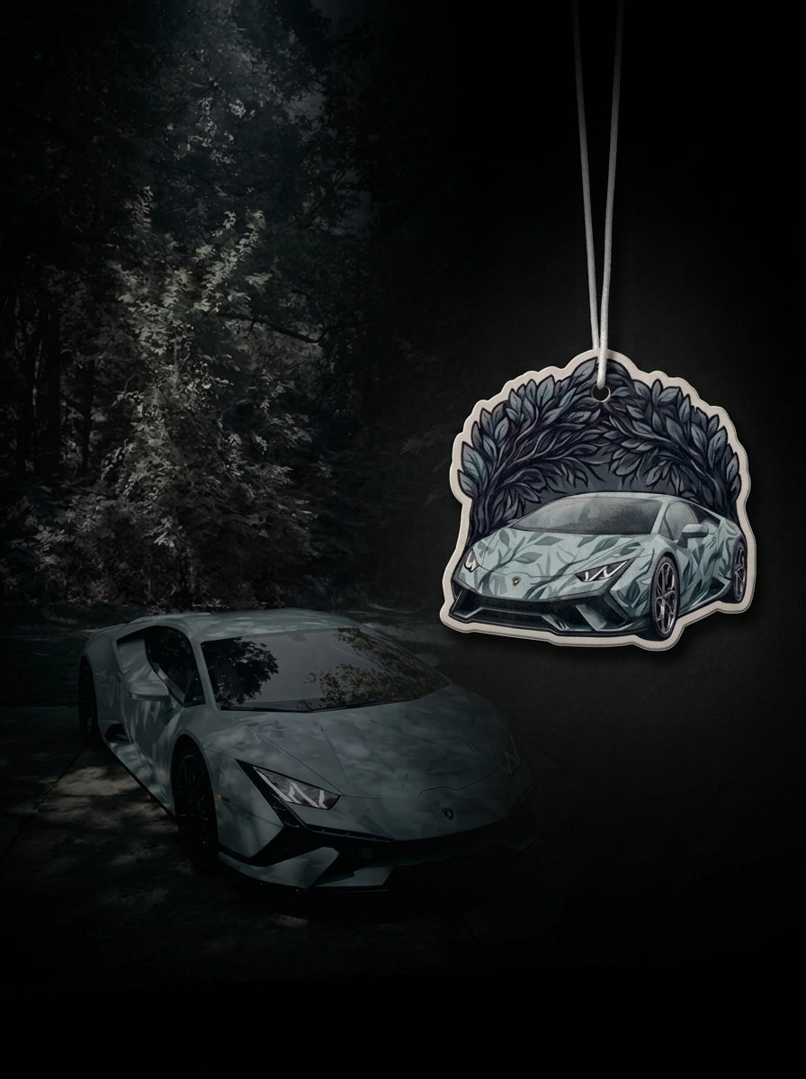 A car-shaped air freshener hanging from a string against a dark forest background, with a Lamborghini sports car illustration printed on the front.