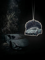 A car-shaped air freshener hanging from a string against a dark forest background, with a Lamborghini sports car illustration printed on the front.