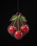 A cherry-shaped hanging ornament or plaque with three glossy red cherries, green leaves, and stems on a dark brown background, suspended by a black cord against a black backdrop.
