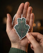 A person holds a die-cut car air freshener shaped like a pine branch in their hand. The product features a detailed green pine branch design with a white border and a white hanging loop, shown against a dark, blurred background.