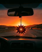 A star-shaped car air freshener hangs from a rearview mirror in a car, backlit by a bright orange sunset over a highway.
