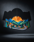 A colorful mountain landscape ornament with a winding road, pine trees, and a sunburst, hanging against a dark gray background with a cast shadow.