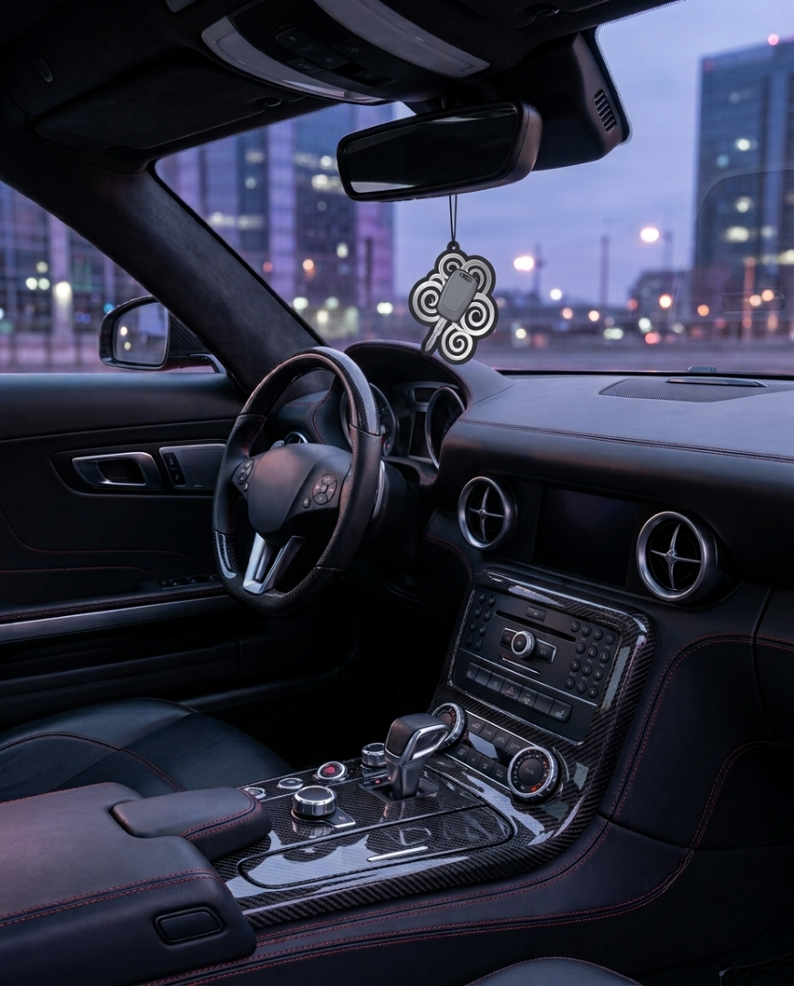 A Mercedes-Benz car interior with a hanging silver and black decorative ornament in the center console, shown at dusk with a blurred city skyline outside the windshield.