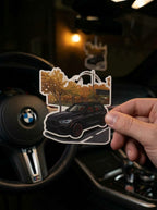 A hand holds a die-cut sticker featuring a black BMW SUV with autumn trees and a street sign, set against a blurred car interior with a visible BMW steering wheel.