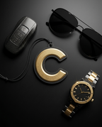 A luxury flat-lay arrangement on a dark background featuring a gold and black letter 'C' keychain, a black carbon fiber car key case, black sunglasses, and a gold and silver metal link watch.