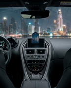 A car interior dashboard view at night with a hanging blue oval air freshener or diffuser centered above the console, in front of a city skyline seen through the windshield.