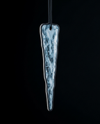 An elongated, tapered glass ornament with a textured, icy blue and white pattern inside a clear glass body, suspended by a black cord against a solid black background.