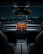 A decorative car air freshener or dashboard ornament hanging in the center console of a car interior at night, with a city skyline visible through the windshield. The ornament has a glowing amber and orange design with a stylized emblem in the middle.