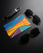 A colorful, geometric-shaped card holder or wallet with a vibrant abstract sunset pattern, styled in a flat-lay arrangement with a black leather keychain and two pairs of black sunglasses on a dark gray background.