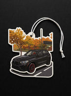 A die-cut car-shaped air freshener or ornament featuring a black BMW SUV in front of an autumn tree scene, with a white hanging cord, shown on a black background.