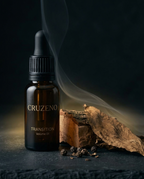 A dark amber glass dropper bottle labeled CRUZENO TRANSITION Volume 87, styled on a dark surface with a dried leaf, wood piece, and scattered seeds under moody lighting with smoke.