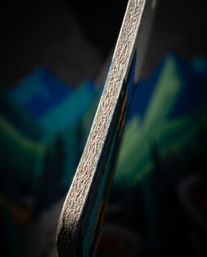 Extreme close-up of a skateboard deck's side edge, showing the textured grip tape and layered wood construction, with a blurred multicolor mountain graphic in the background.