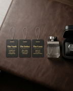 Three black leather fragrance gift tags labeled 'The Vault', 'The Ember', and 'The Coast' with gold text, displayed next to a clear glass perfume bottle and an open black watch box on a brown leather surface.