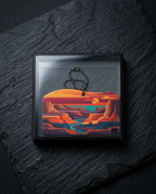 A square framed art print or pendant featuring a stylized desert canyon landscape in orange, red, teal, and purple tones, displayed on a dark stone surface with a black beaded keychain hanging from the top.