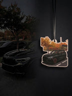 A die-cut car air freshener featuring a black BMW SUV with orange autumn trees, shown hanging against a dark, moody background with a blurred black car and trees.