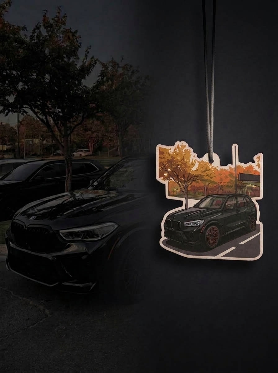 A die-cut car air freshener featuring a black BMW SUV with orange autumn trees, shown hanging against a dark, moody background with a blurred black car and trees.