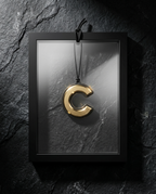 A polished gold letter 'C' pendant hanging from a black cord inside a black frame, set against a dark textured slate background.