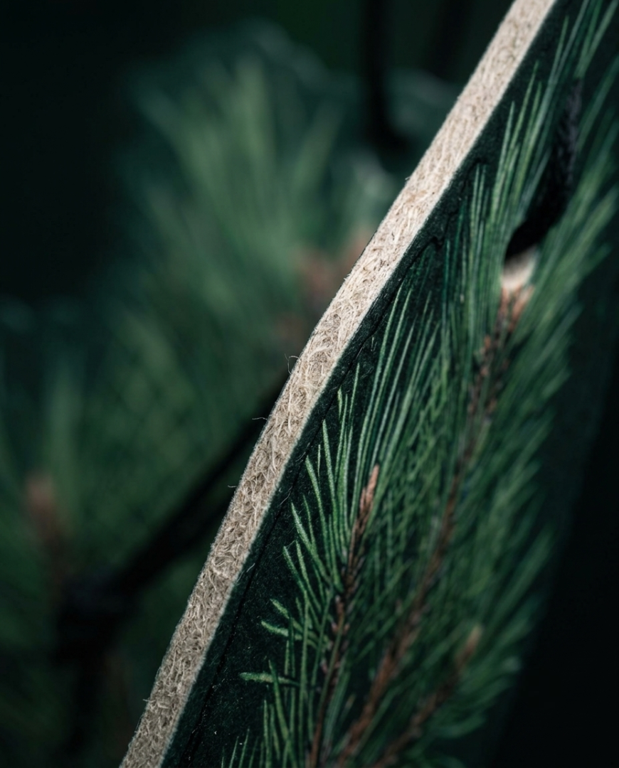 Close-up of a decorative wooden board or sign featuring a dark green pine needle and branch pattern, with a natural wood edge visible against a blurred dark green background.