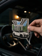 A hand holds a die-cut car air freshener against a blurred car interior background. The air freshener features a white sports car under a modern bridge with green trees and blue sky details.