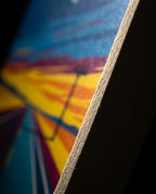 Close-up of a colorful printed panel or artwork edge, showing a blurred abstract sunset-like design in blue, yellow, orange, and purple with a visible wood or cardboard edge in the foreground against a dark background.