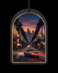 An arched black car air freshener ornament with a gold inner border, featuring a printed sunset cityscape of a palm-lined street with a convertible car and skyscrapers, shown against a black background.