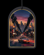 An arched black car air freshener ornament with a gold inner border, featuring a printed sunset cityscape of a palm-lined street with a convertible car and skyscrapers, shown against a black background.