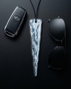 A black car key fob, black sunglasses, and a long sculptural pendant with a white and gray marbled, ice-like pattern, arranged on a dark black background.