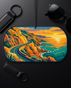 A rectangular car air freshener or hanging ornament with rounded corners, featuring a stylized coastal road scene in warm orange, yellow, teal, and green tones. It is photographed on a dark surface with sunglasses, keychains, and a cord visible around it.
